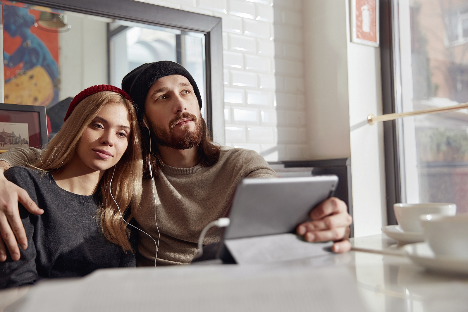 Young thoughtful european couple in headphones watching something on digital tablet in modern cafe.