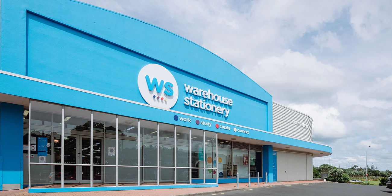 A large, blue Warehouse Stationery store with glass windows and the company logo above the entrance on a cloudy day.