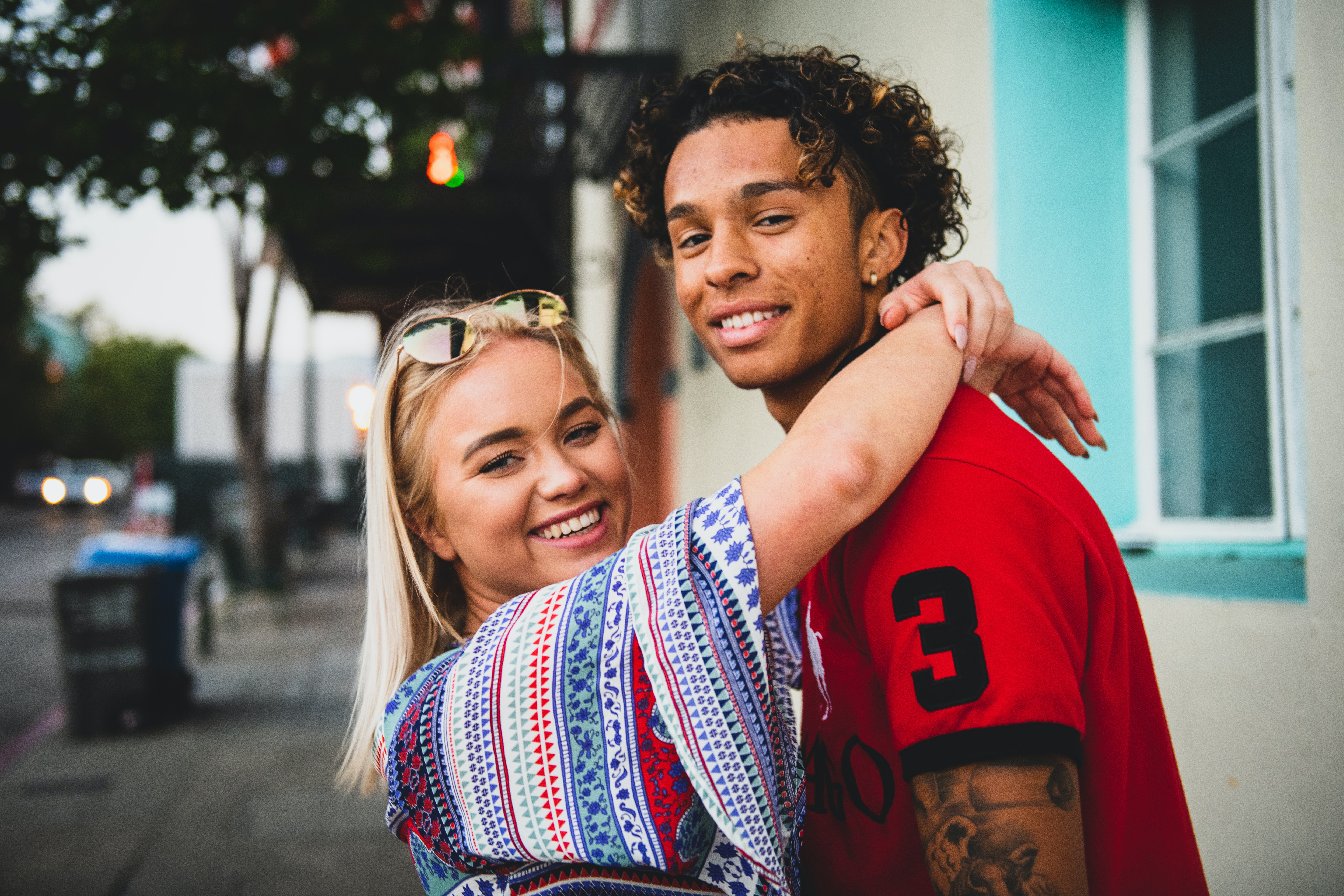 Image of a blond haired woman wearing a summer dress and an attractive mutliracial young man wearing a polo shirt. Both are holding eachother and smiling looking at the camera in the street.