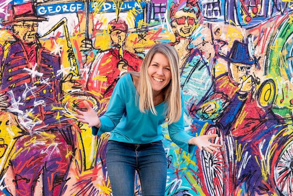 Image of Sally Dear, founder of Ducky Zebra, smiling with her arms stretched out infront of a brightly painted wall