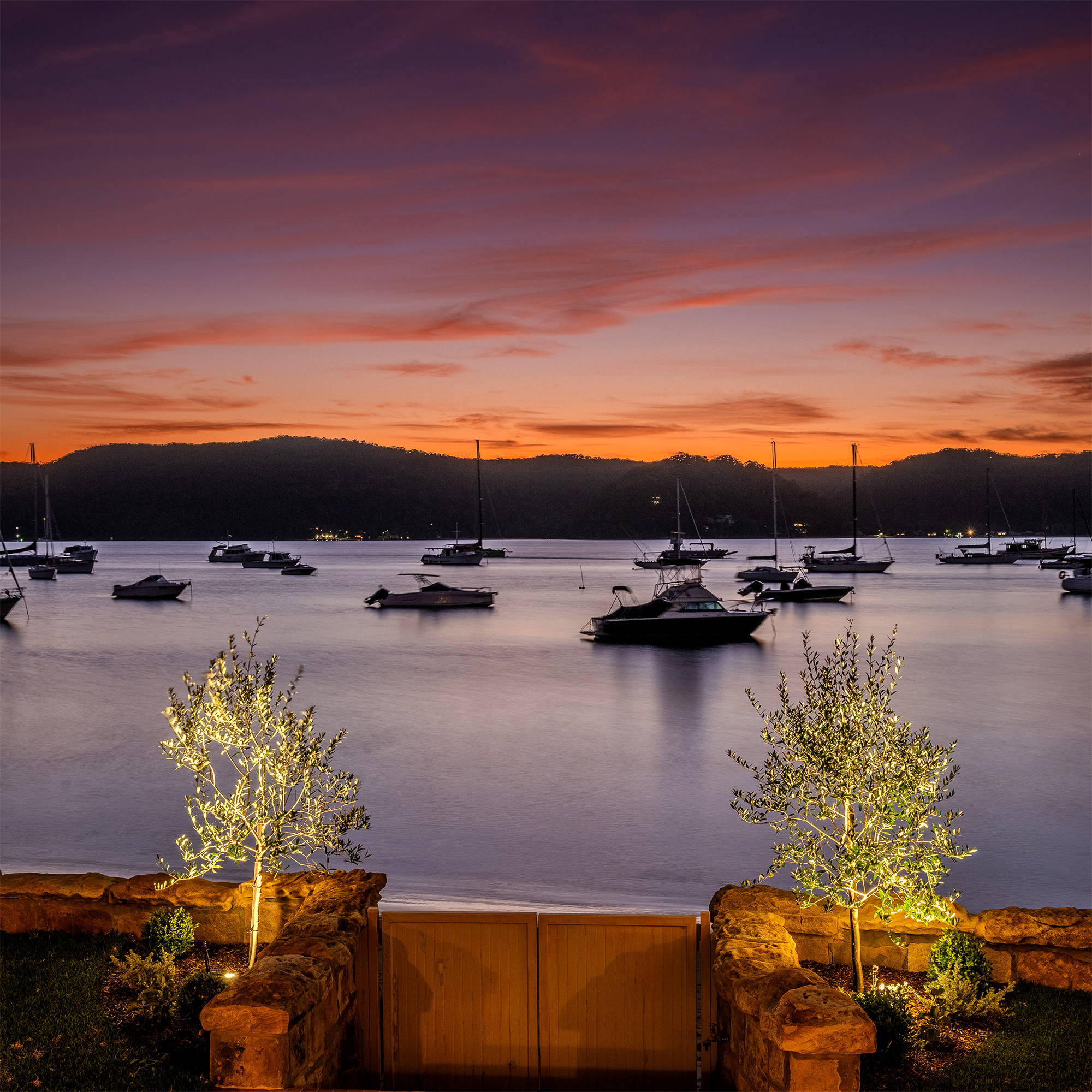 Havit Lighting - Outdoor Lighting by Havit - Inground Light beaming, uplighting trees at the backyard jetty at dusk time, with mountains and an orange sky in the horizon