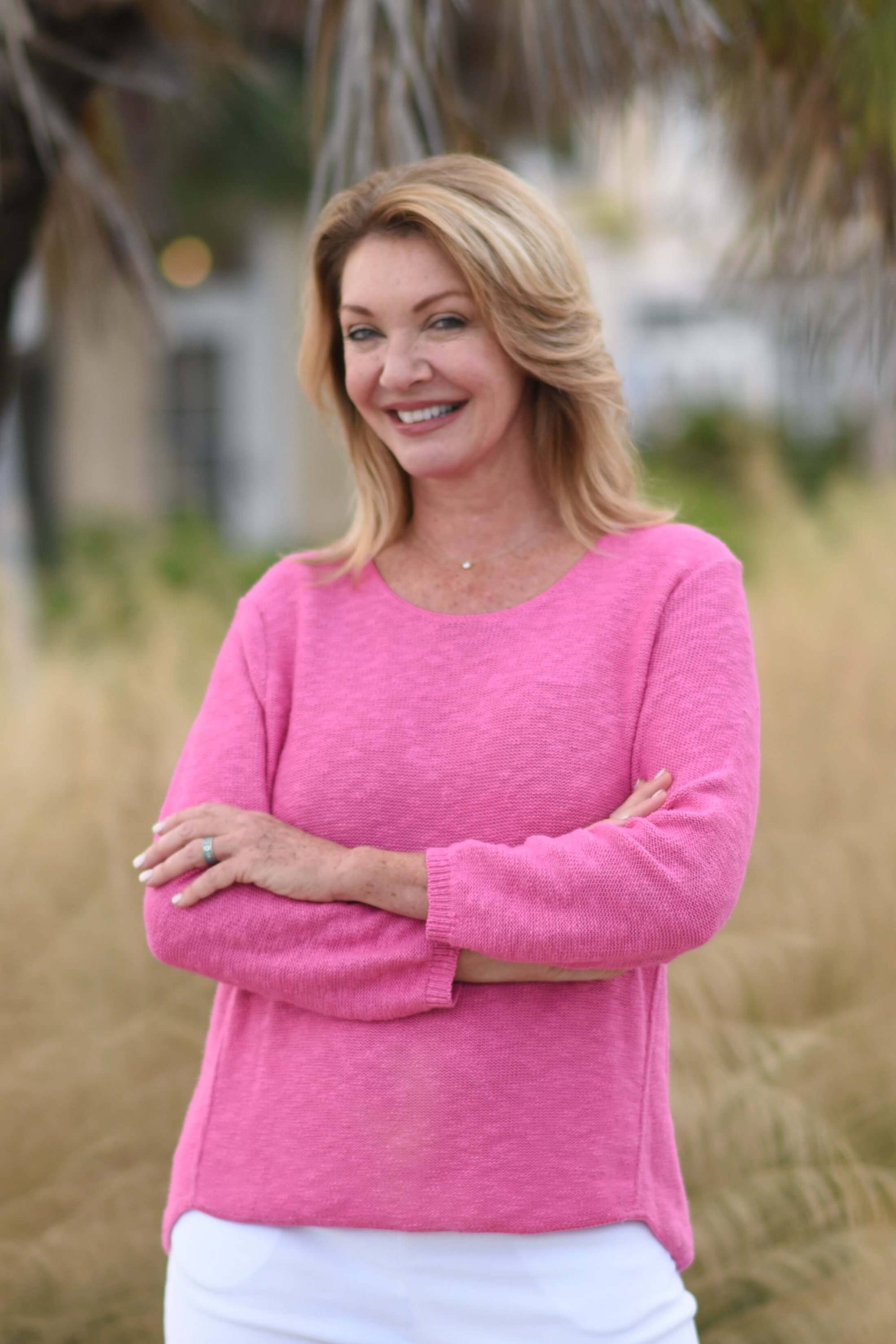 A woman wearing a pink sweater and white pants crosses her arms in front of her chest and she stands in a grassy area