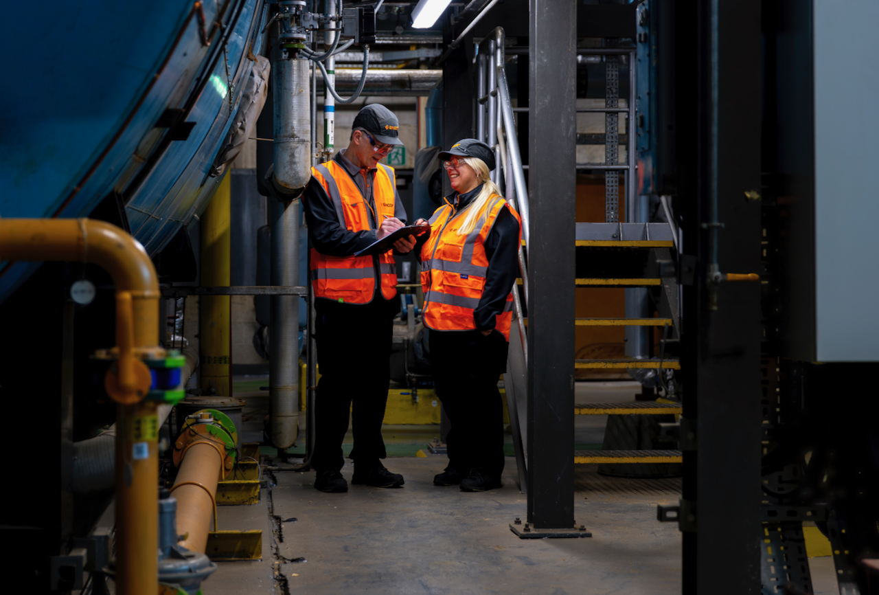 Two workers wearing orange safety vests and hats stand in an industrial facility, talking and looking at a tablet. They are surrounded by pipes, machinery, and metal structures. The environment appears clean and organized.