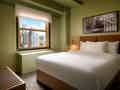 Modernes Hotelzimmer mit Kingsize-Bett, hellgrünen Wänden und einem Fenster mit Blick auf die Skyline.