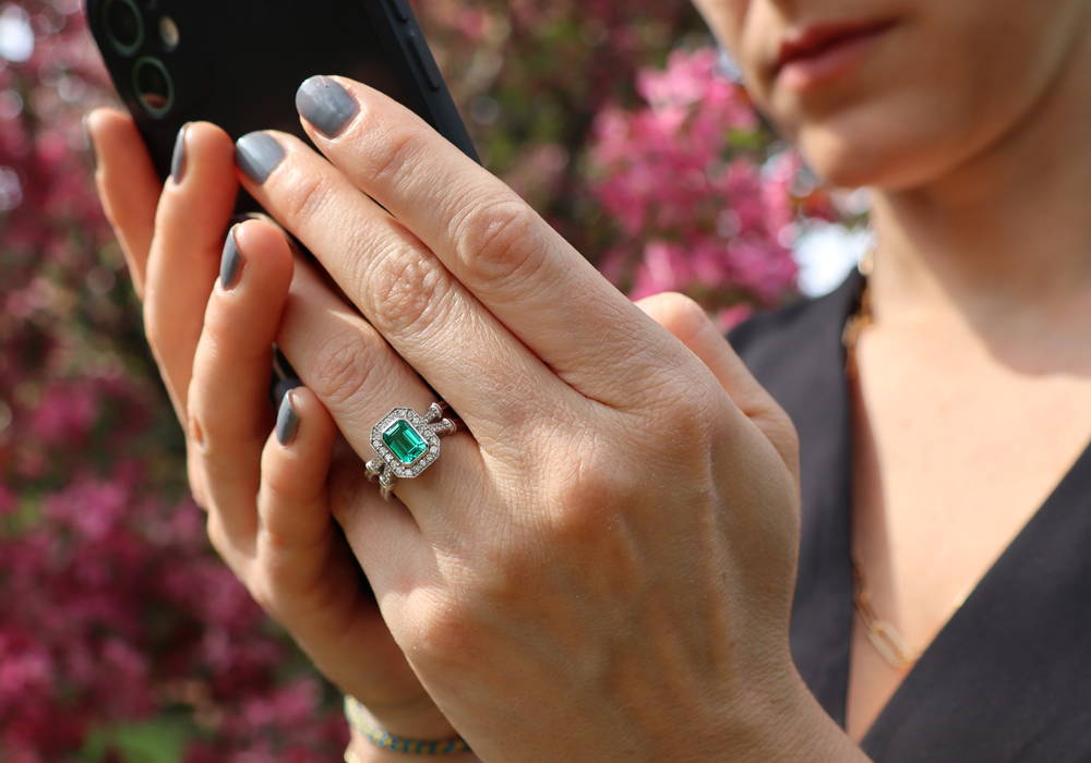 Sublime bague pour femme avec pierre précieuse entourée de diamants en halo. Corps de bague double avec pavés de diamants sur main qui tient un portable.