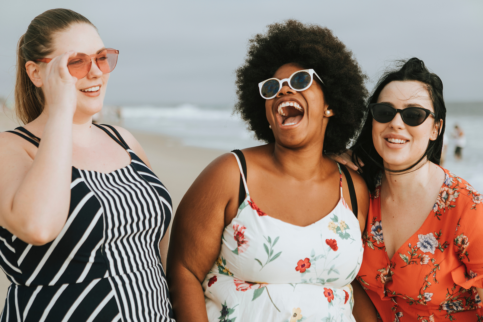 Image of 3 curvy and attractive friends of mixed ethnicities, all wearing floral dresses and sunglasses , walking side by side laughing together.