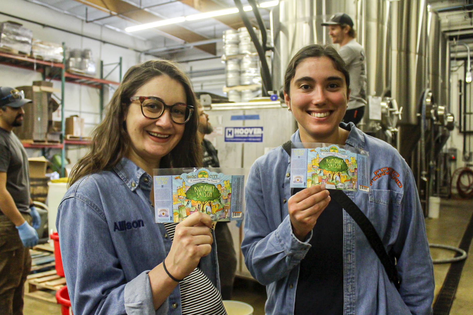 Two members of Food Forward holding Kumquat Saison beer labels at the Smog City Brewery