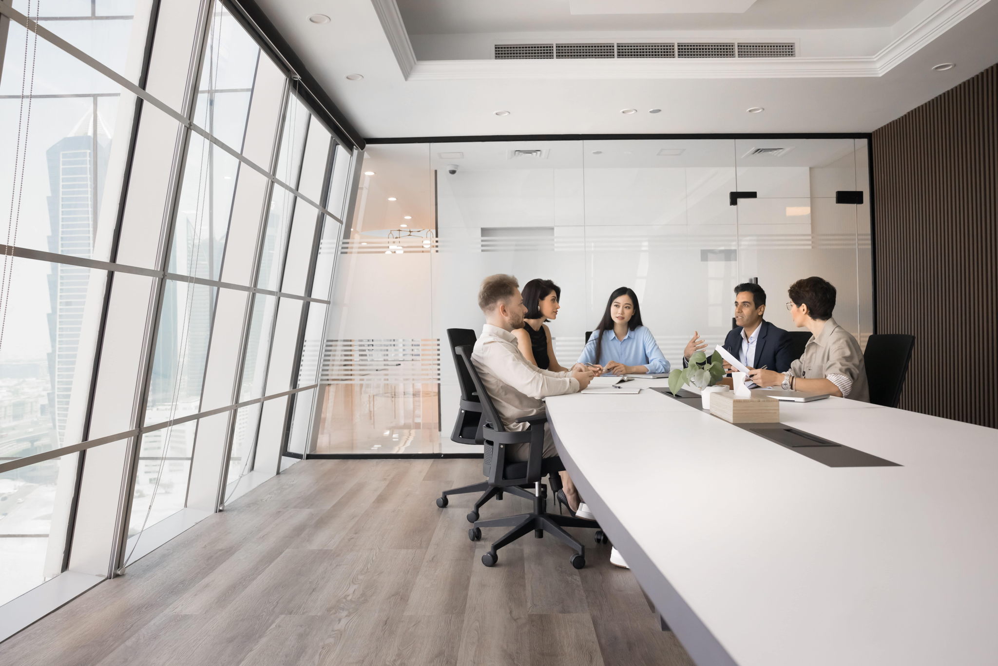 Five people are seated around a long conference table in a modern office, designed through our workplace services, engaged in discussion. Large windows provide a view of tall skyscrapers outside. The room features wood flooring and white walls with some decorative elements.