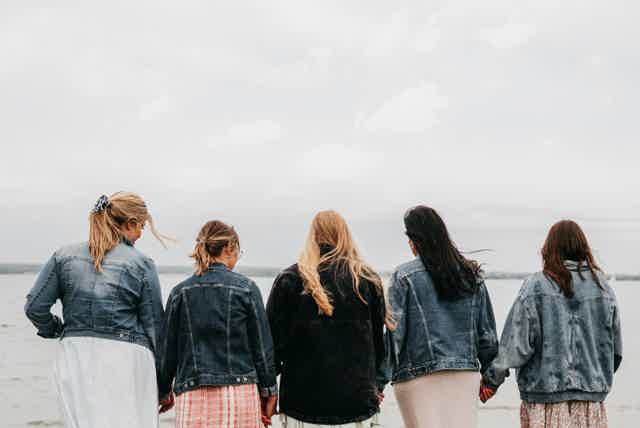 Back view of five women standing hand-in-hand outdoors, facing away from camera in a park.
