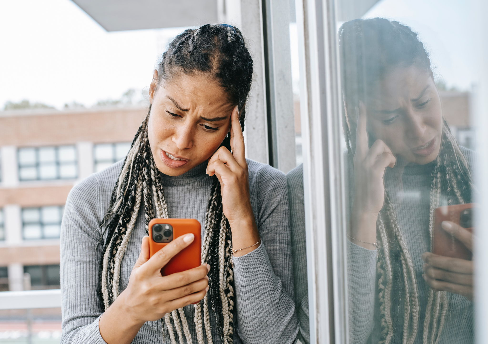 A woman with dreads sits on her patio looking on her phone with a concerned look on her face.