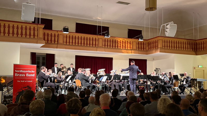 Auf einer Bühne sitzt eine Brass Band in schicken schwarzen Anzügen und gibt ein Konzert. 