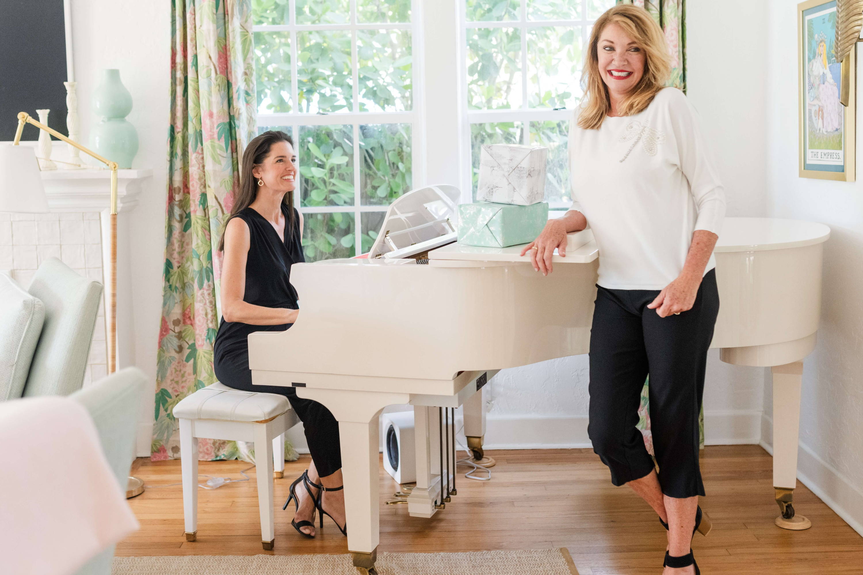 Two women dressed in holiday party attire laugh next to a white piano with wrapped gifts