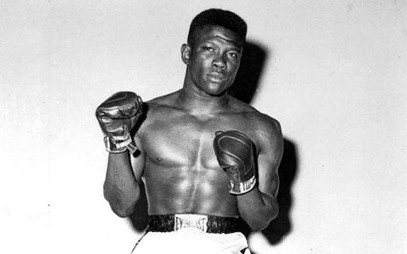 Black and white image of Emile posing for a photo with his gloves and boxing uniform, looking seriously at the camera.