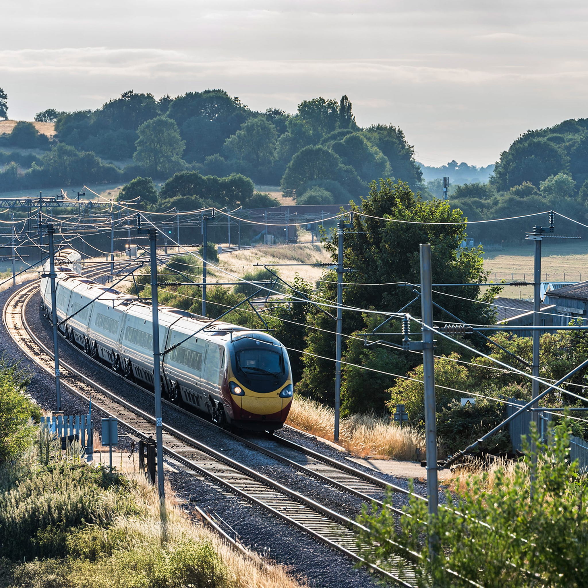 A modern passenger train travels along curved tracks through a green and hilly countryside, with power lines overhead and trees and fields in the background under a partly cloudy sky.
