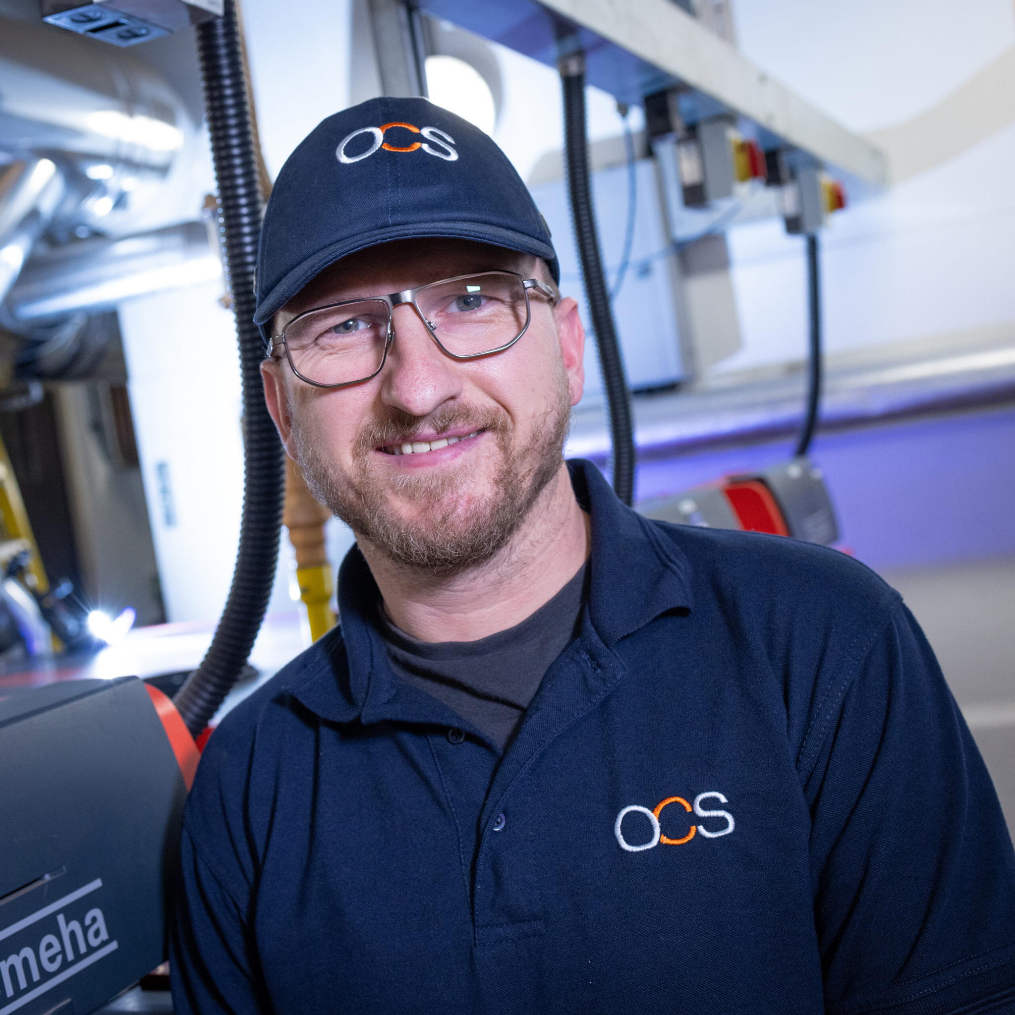 A smiling man wearing glasses, a navy blue OCS cap, and a matching OCS polo shirt stands in an industrial or factory setting with machinery and pipes in the background.