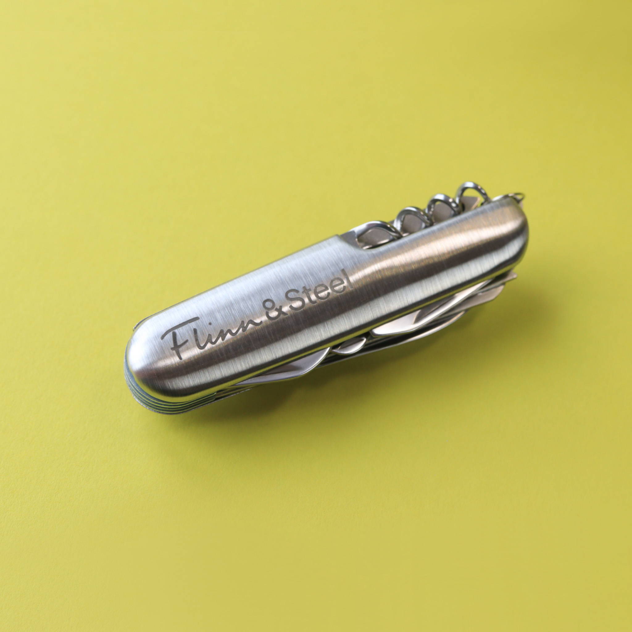 Close up image of a stainless steel multi-tool penknife. The knife is folded in and a Flinn & Steel logo can be seen engraved on the side.