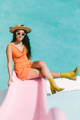 a woman wearing a straw hat, mustard boots, and an orange romper sits on a pink slide wearing sunglasses for "poolside pride: a slim aarons-inspired photo campaign."