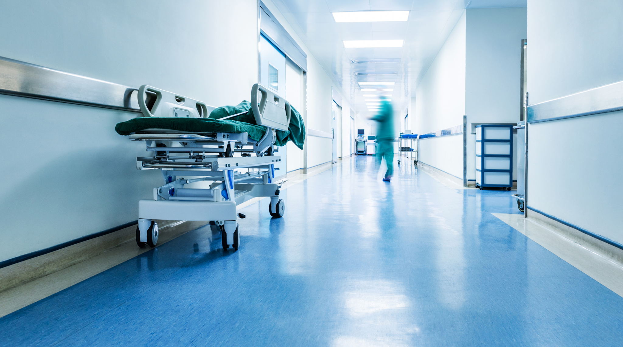 A hospital hallway with blue floors, an empty stretcher on the left, and a blurred medical professional walking in the distance under bright lights.