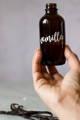 A hand holds up a brown glass bottle labeled 'Vanilla' with vanilla beans sitting on the counter