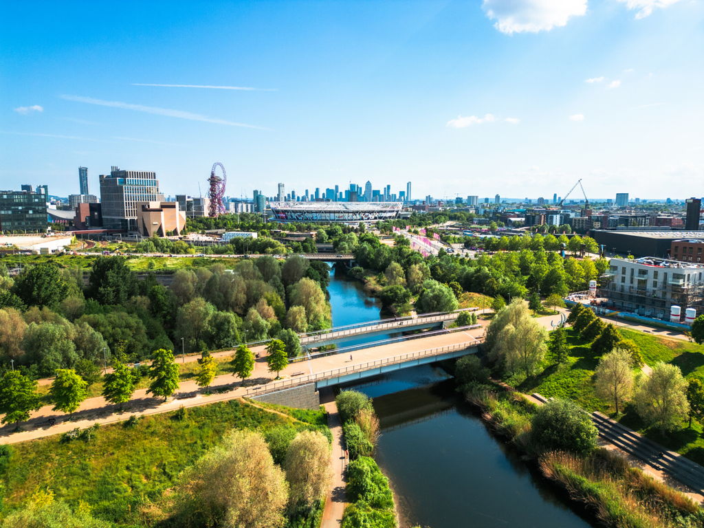 A vibrant cityscape featuring lush green parks, a winding river with footbridges, and modern buildings under a bright blue sky with scattered clouds. The skyline includes tall structures in the distance.