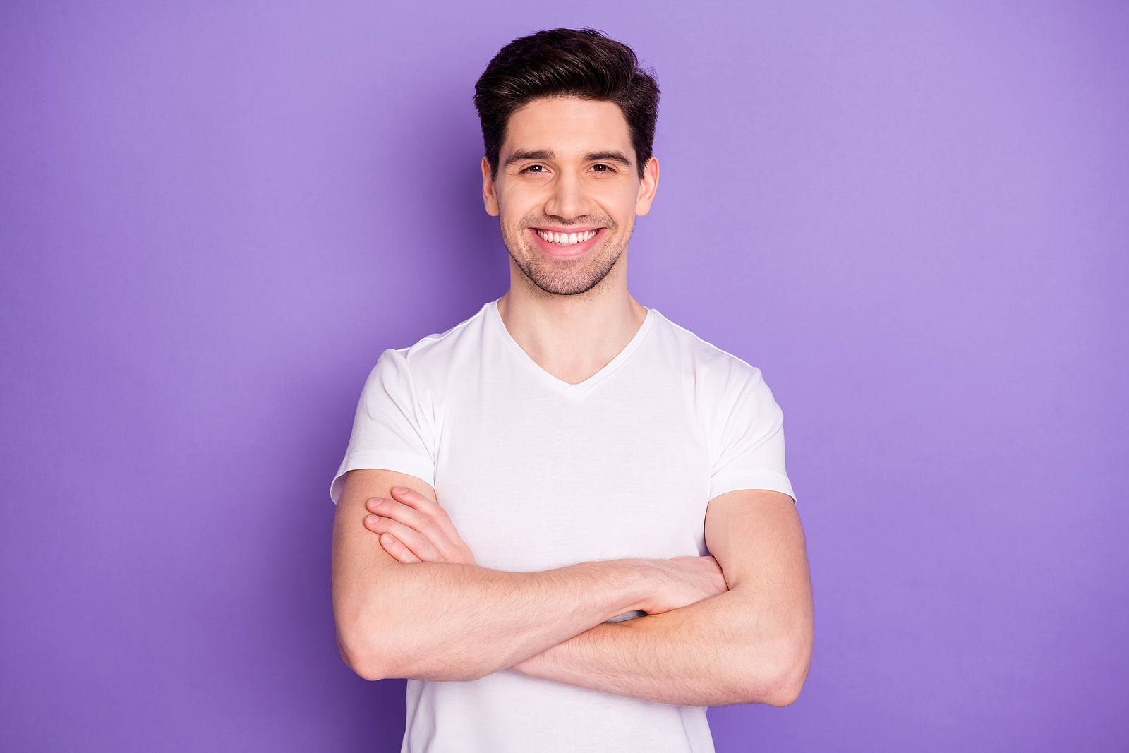 A confident man wearing a white t shirt crosses his arms and smiles against a purple background.