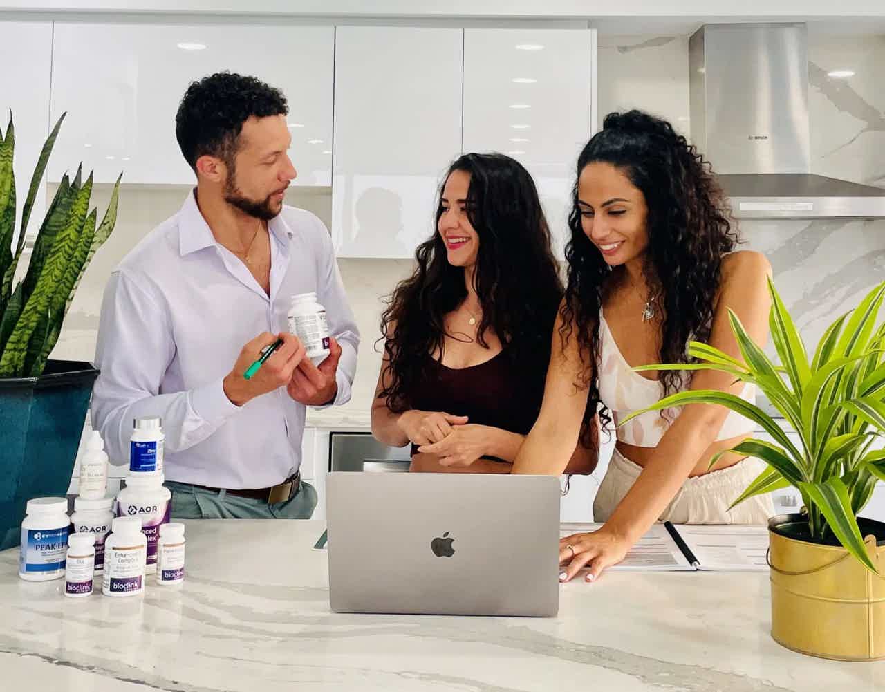 Three people discussing products at a kitchen counter with a laptop and houseplants.