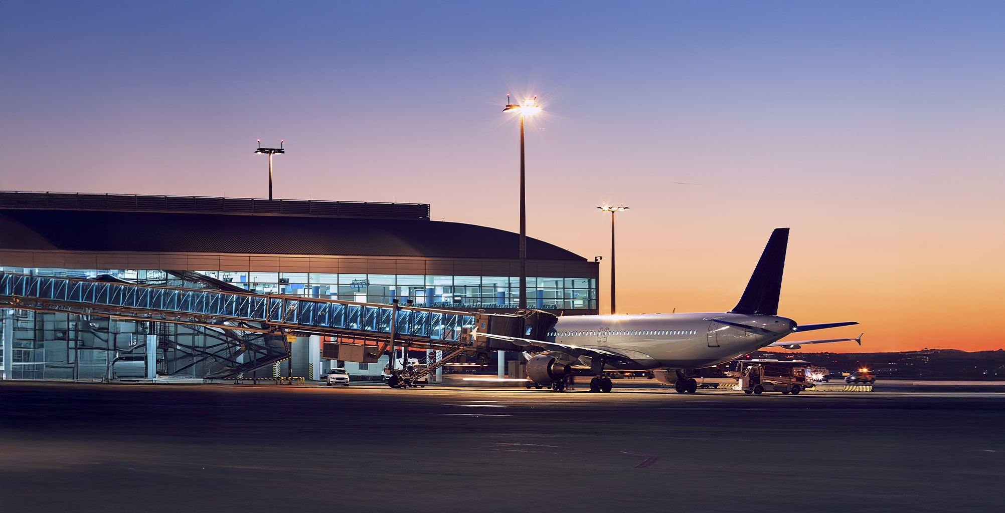 A passenger airplane is parked at an airport gate at sunset, with a boarding bridge connected and airport vehicles nearby. The terminal building is illuminated in the background.