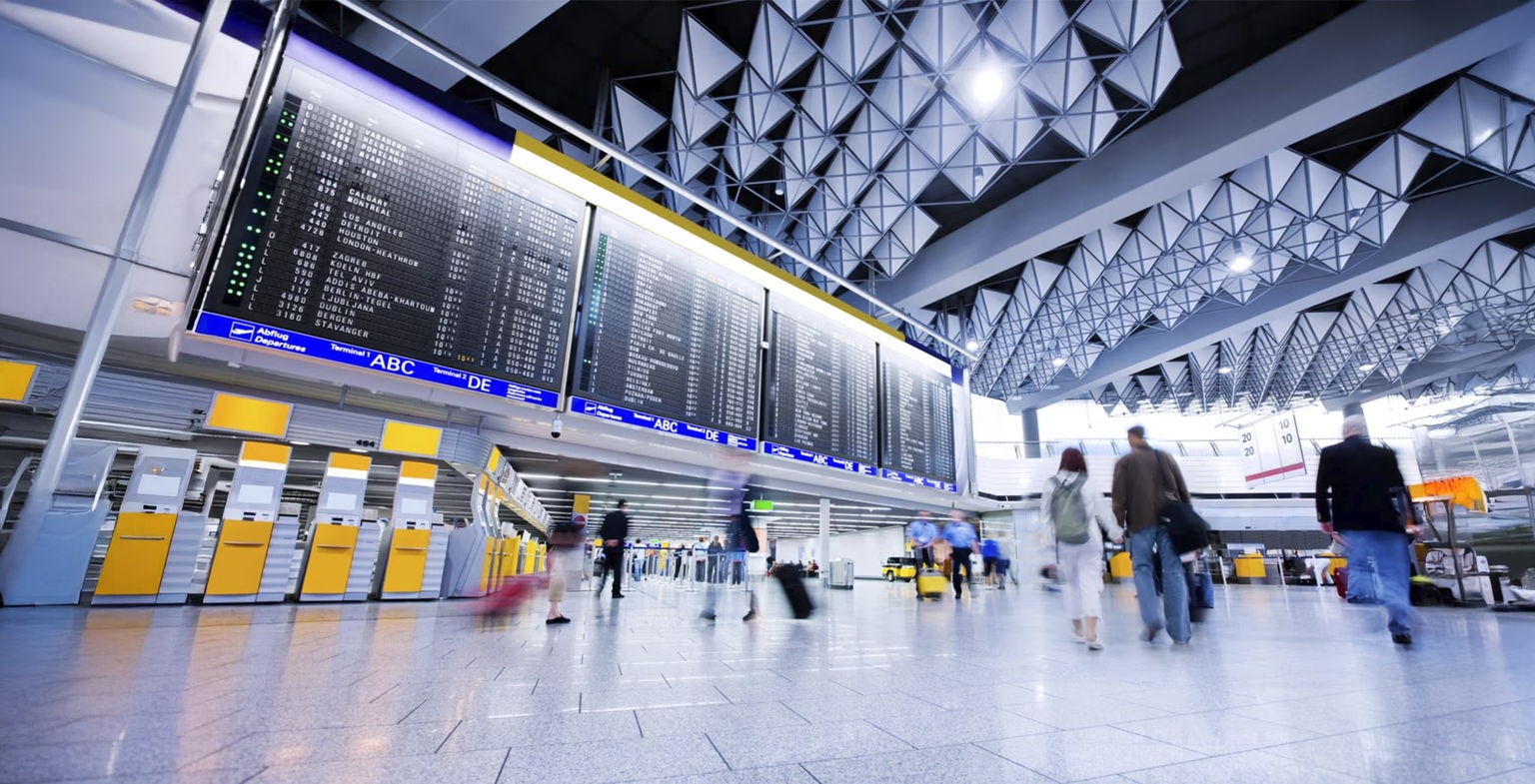 Spacious airport terminal with travelers walking and rolling suitcases, large electronic flight information board above yellow check-in counters, and a high ceiling with geometric patterns.