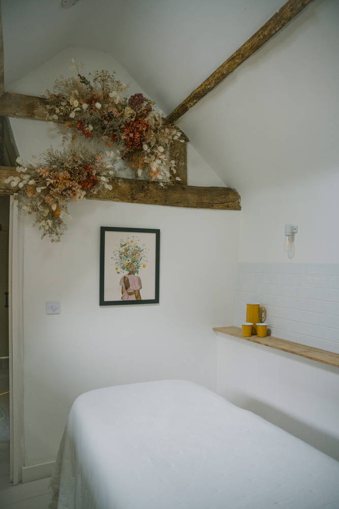 AMLY facial room, white walls, exposed wooden beams, white sheet on a bed, dried flowers on the wall