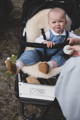 A baby, sat up, facing forward, sat on a sheepskin pram liner. The baby has brown lambskin booties and is in a mountain buggy pram. a mum is to the side of the baby, mostly off screen.