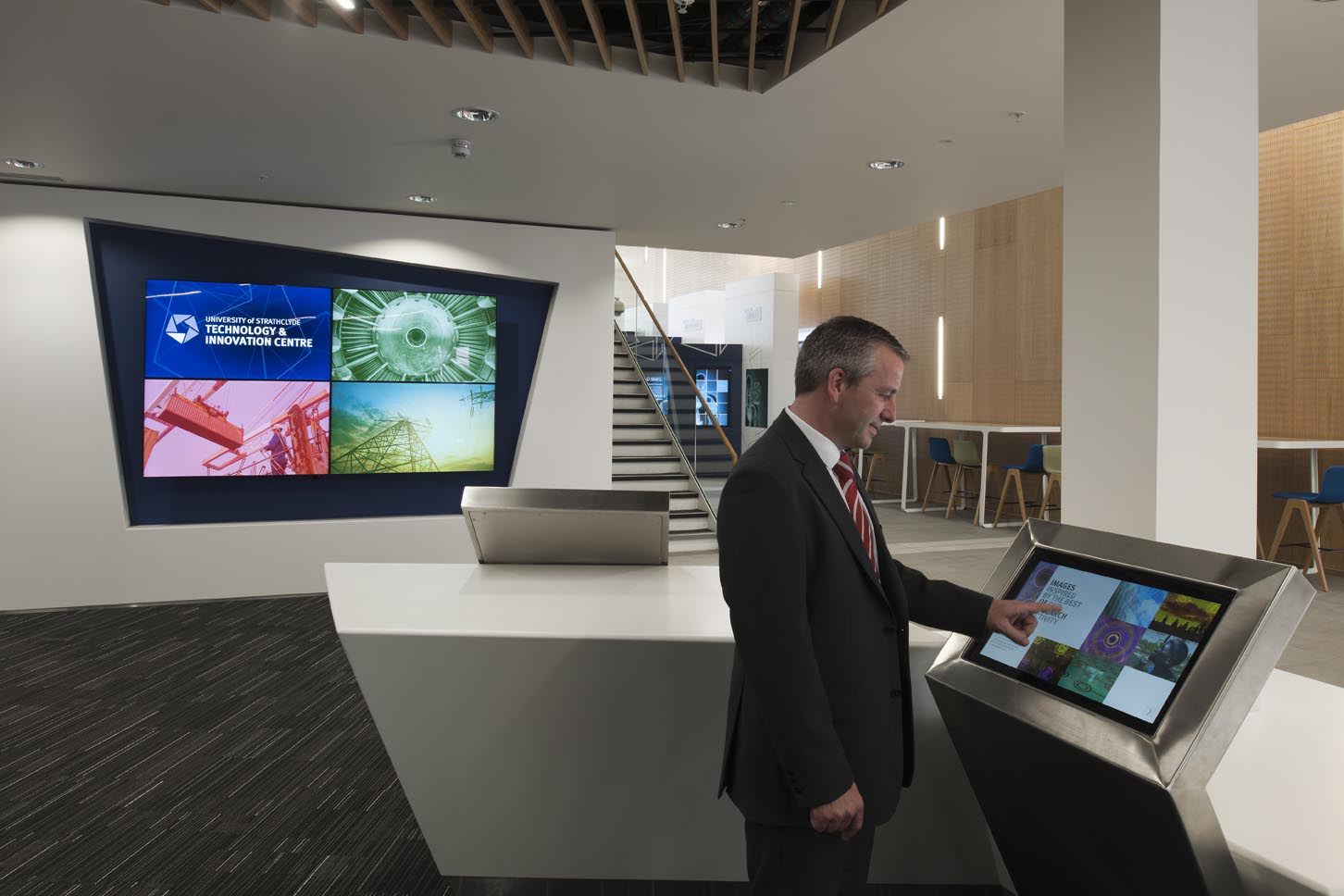 A man in a suit uses a touchscreen kiosk in a modern office lobby with digital display screens on the wall and high tables with blue chairs in the background.