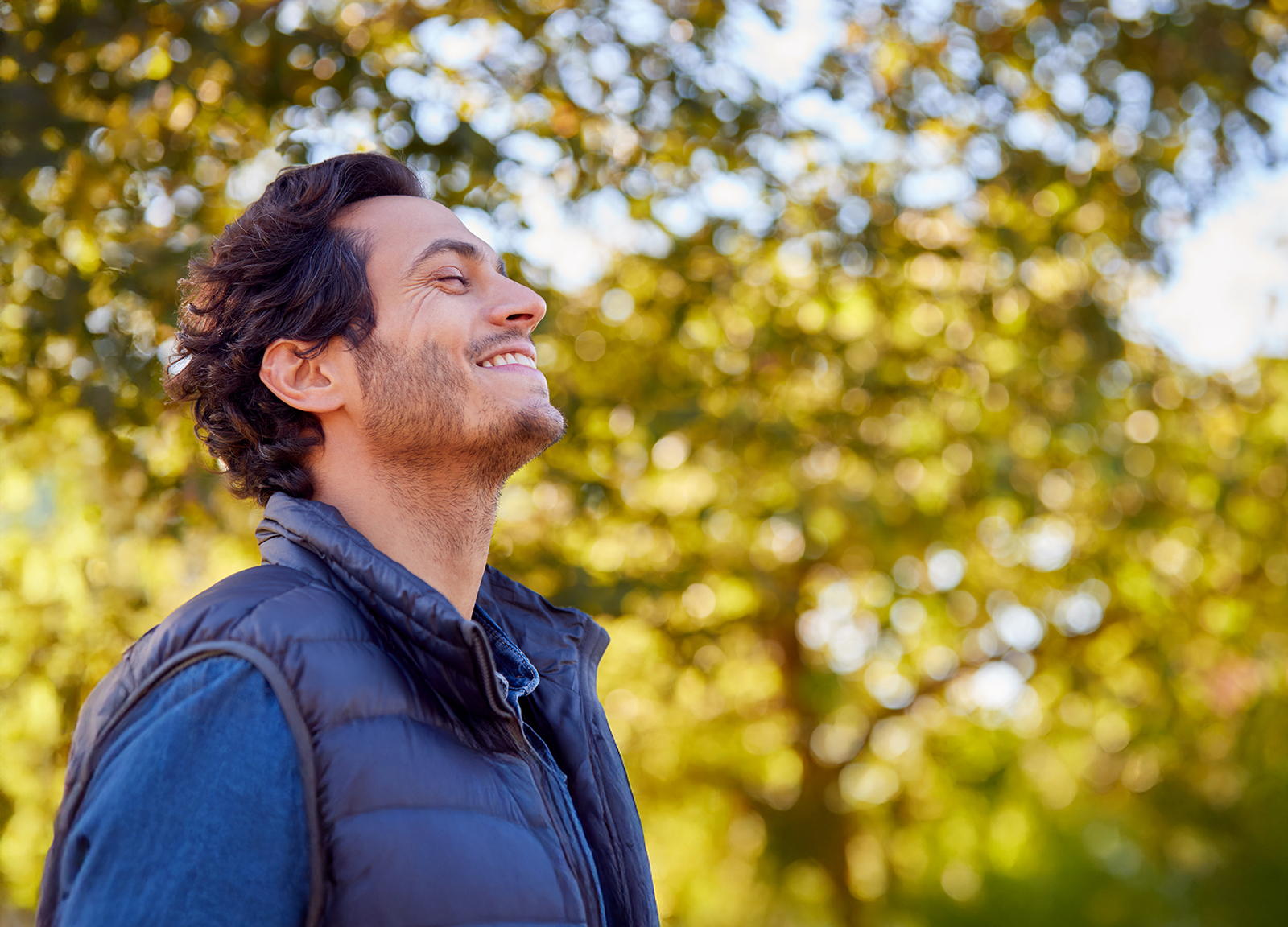 A man with dark hair smiles and tilts his head back while standing outdoors in a sunlit area with green, leafy trees in the background. He wears a blue shirt and a black puffer vest.