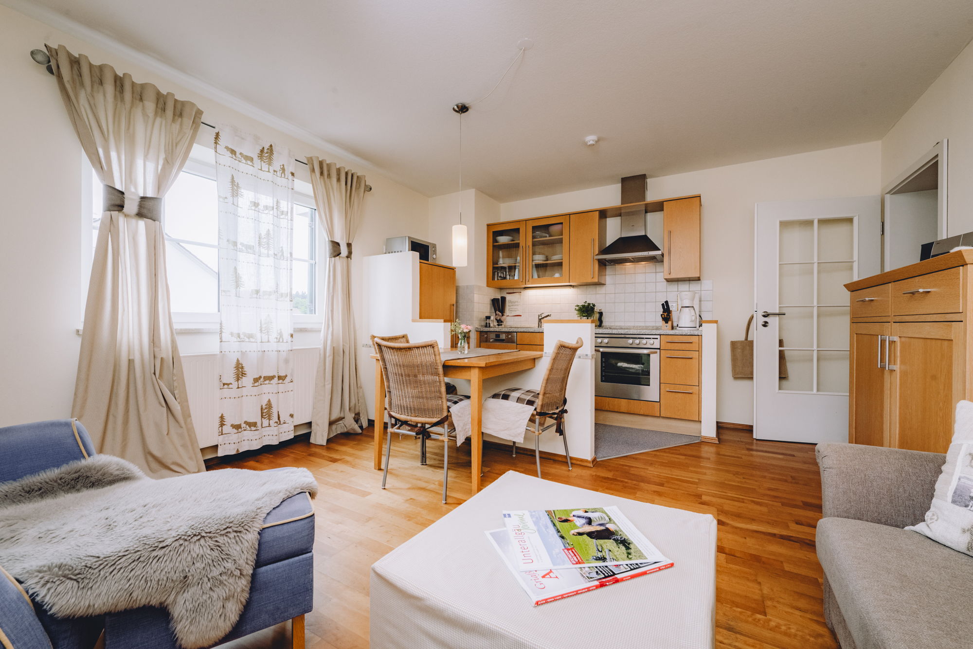 Im Vordergrund ein Wohnraum mit blauen und grauen Sitzmöglichkeiten, im Hintergrund ein hölzerner Essbereich und eine mit Holz verkleidete Küche.