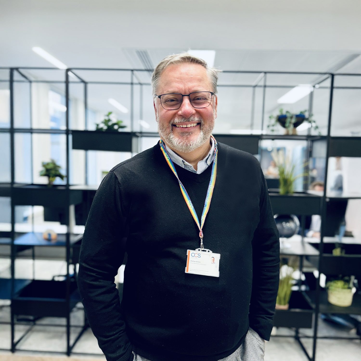 A smiling man wearing glasses, a black sweater, and a lanyard with an ID badge stands in an office with black shelving and potted plants in the background.