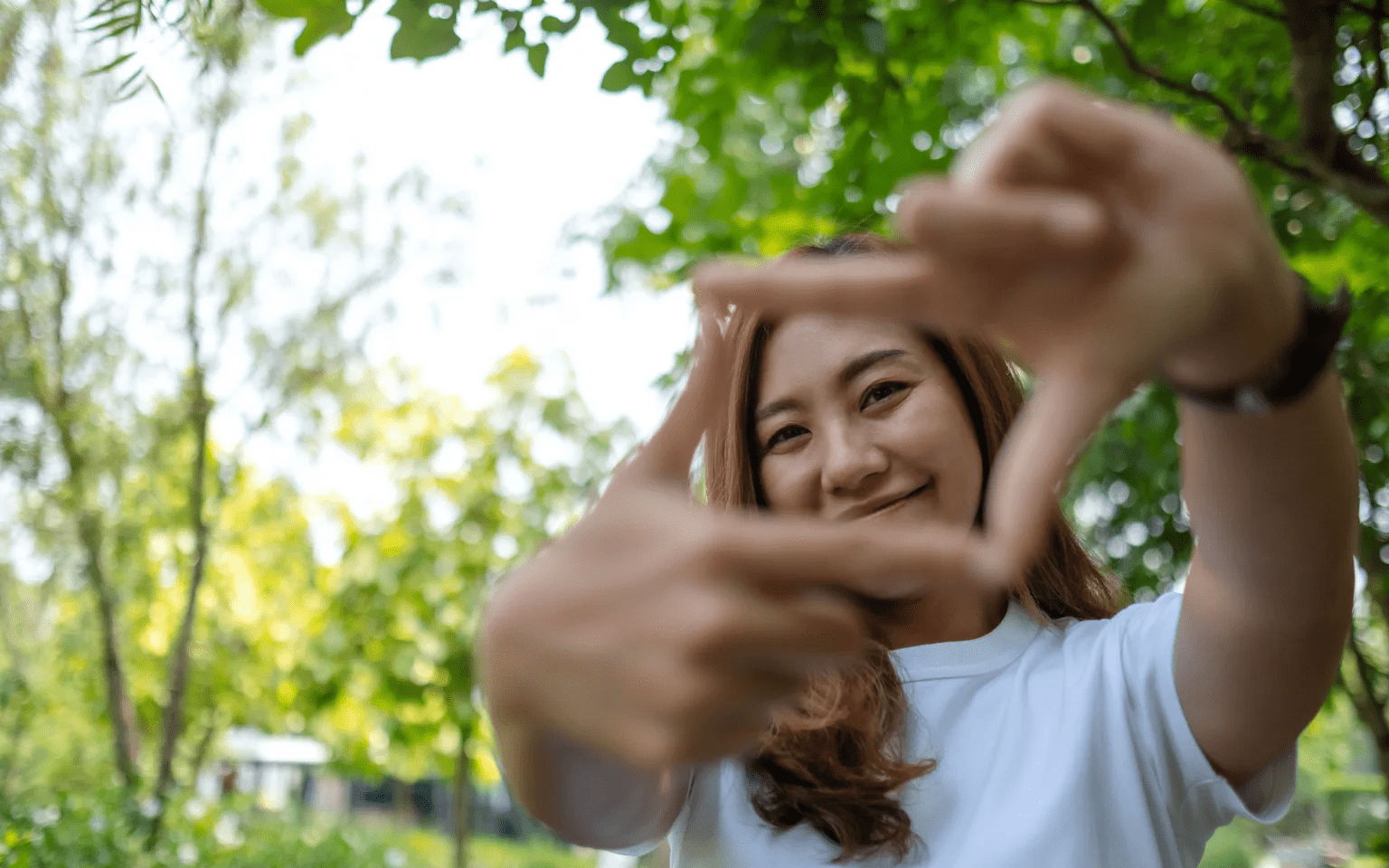 A young woman in a white shirt smiling outdoors, framing her face with her fingers to form a rectangle. Green trees and sunlight are visible in the background.