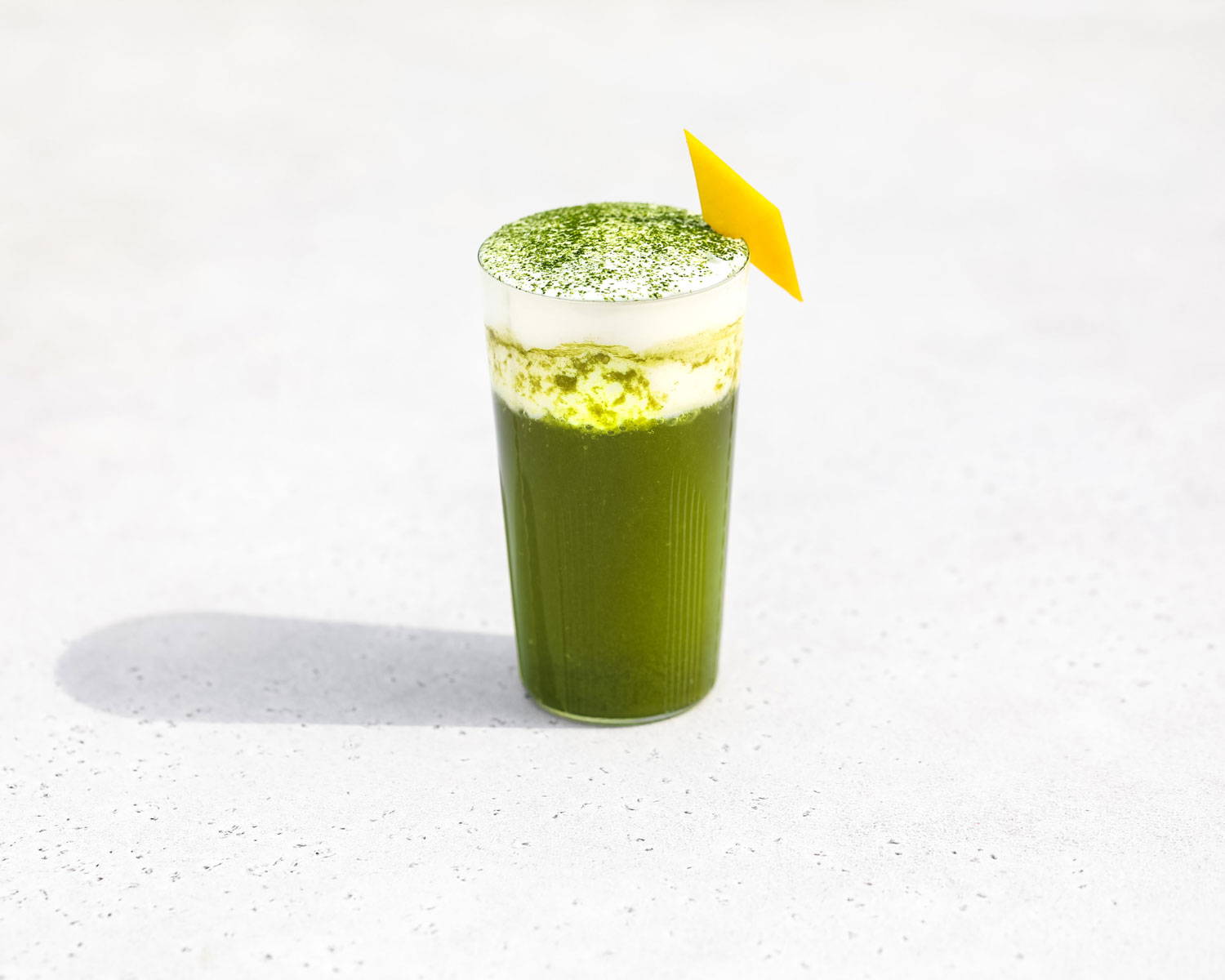 A glass of bright green, sparkling yuzu matcha topped with cream and sprinkled with matcha powder.