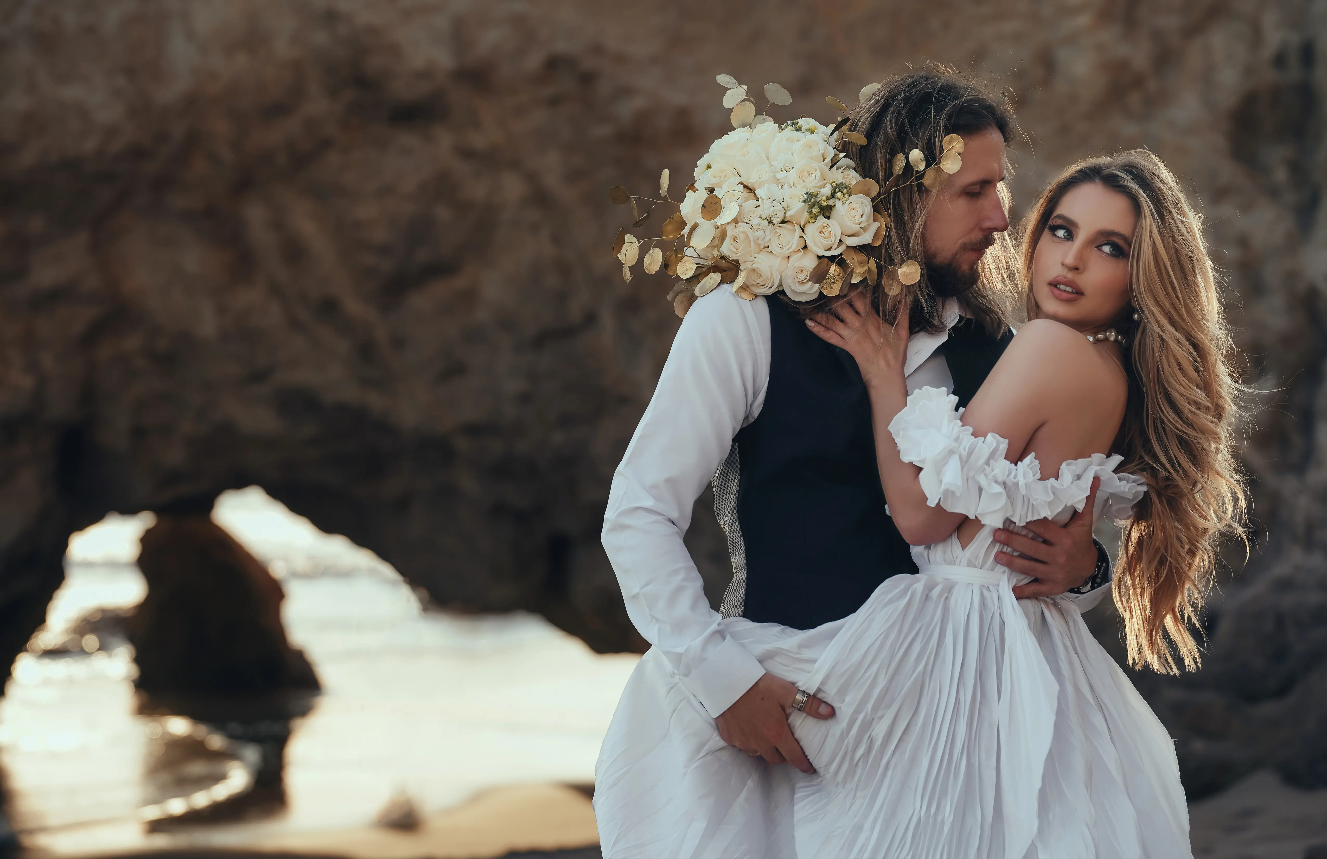 Un moment enchanteur de deux jeunes mariés se tenant sur une plage californienne, avec leur robe blanche et leur gilet noir.