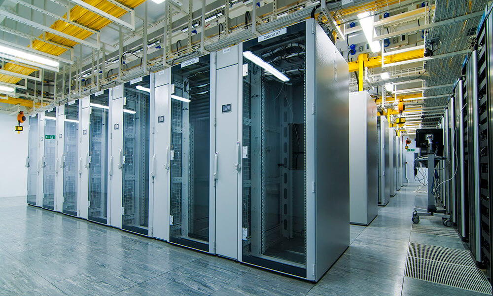 A row of tall, glass-fronted server racks lines a brightly lit, modern data center with a reflective floor and exposed overhead cables.