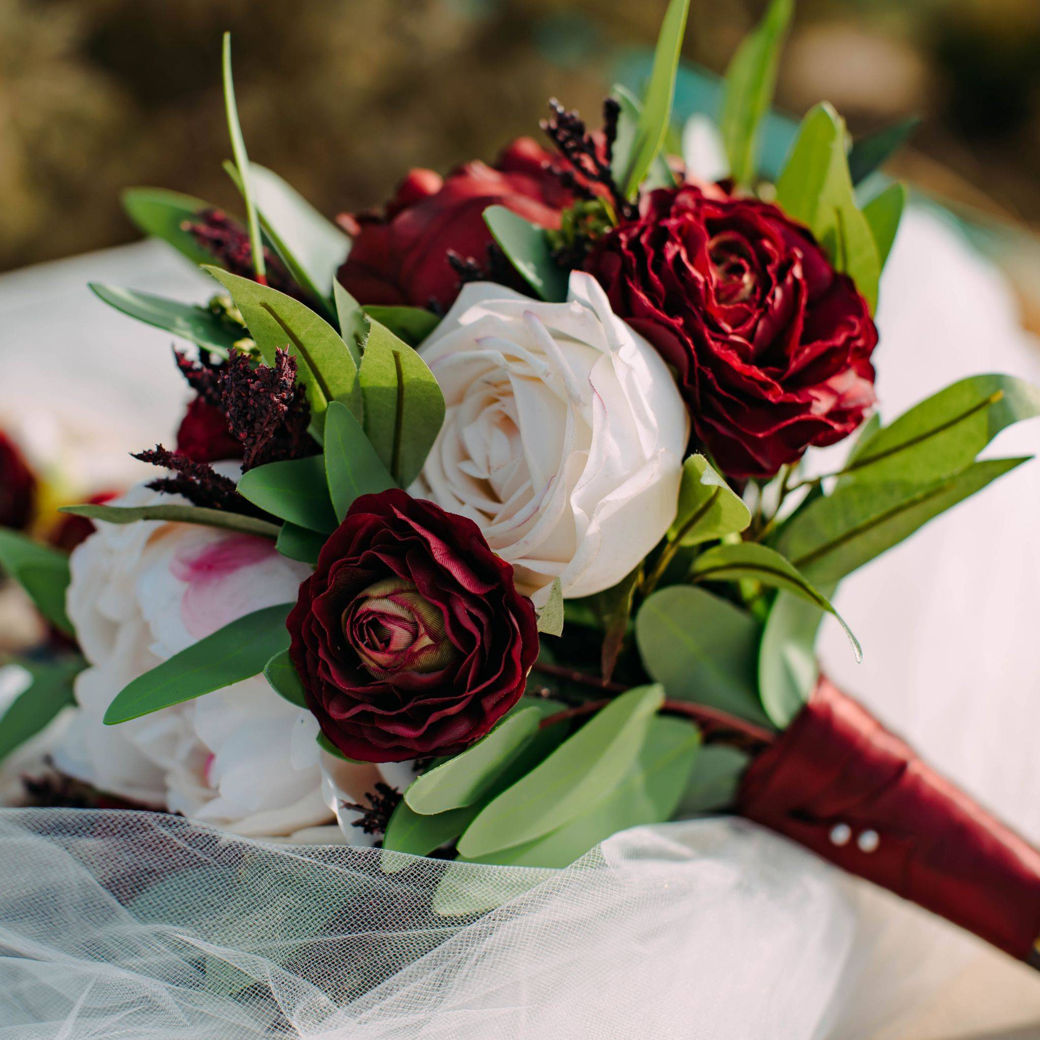 A bouquet made of burgundy ranunculus, blush peonies, and blush cabbage roses with ruscus and seeded eucalyptus is wrapped in burgundy ribbon laying on a bride's veil