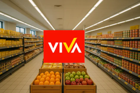 An aisle inside a grocery store filled with shelves stacked with various food products and crates of fresh oranges and apples in the foreground, featuring a Viva logo overlay.
