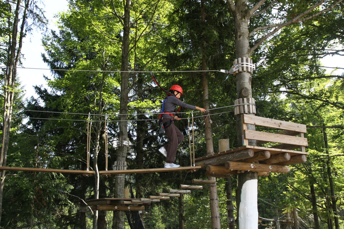 Eine Person in Sicherheitsausrüstung überquert eine Holzbrücke in einem Outdoor-Kletterwald.