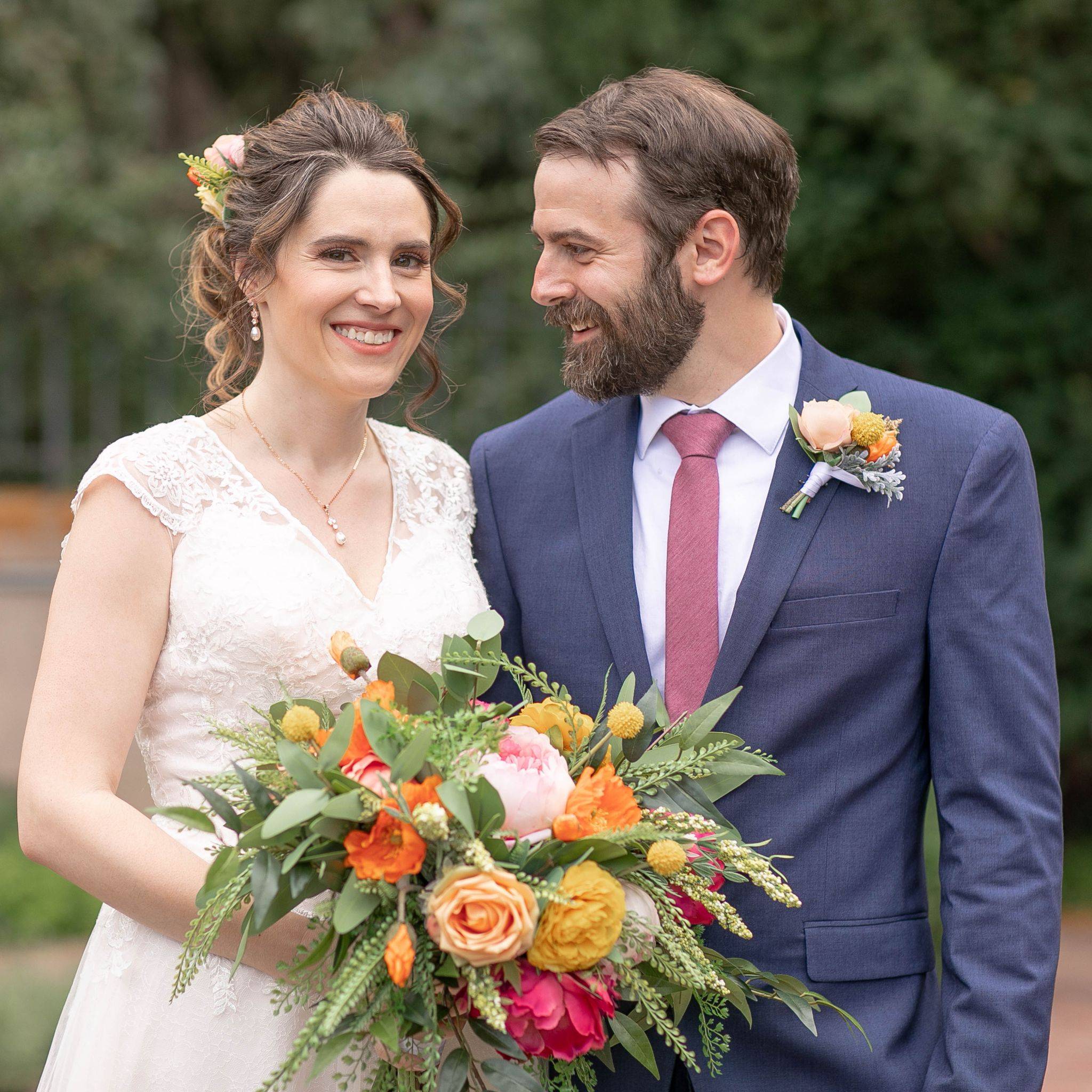 A groom wearing a blue suit with a colorful boutonniere looks at his new bride who is holding a large unstructured bouquet filled with pink peonies, orange roses, yellow ranunculus, eucalyptus and ferns 