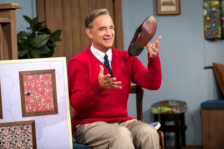 Tom Hanks as Mr. Rogers, throwing a shoe in the air and smiling, sitting in his house.