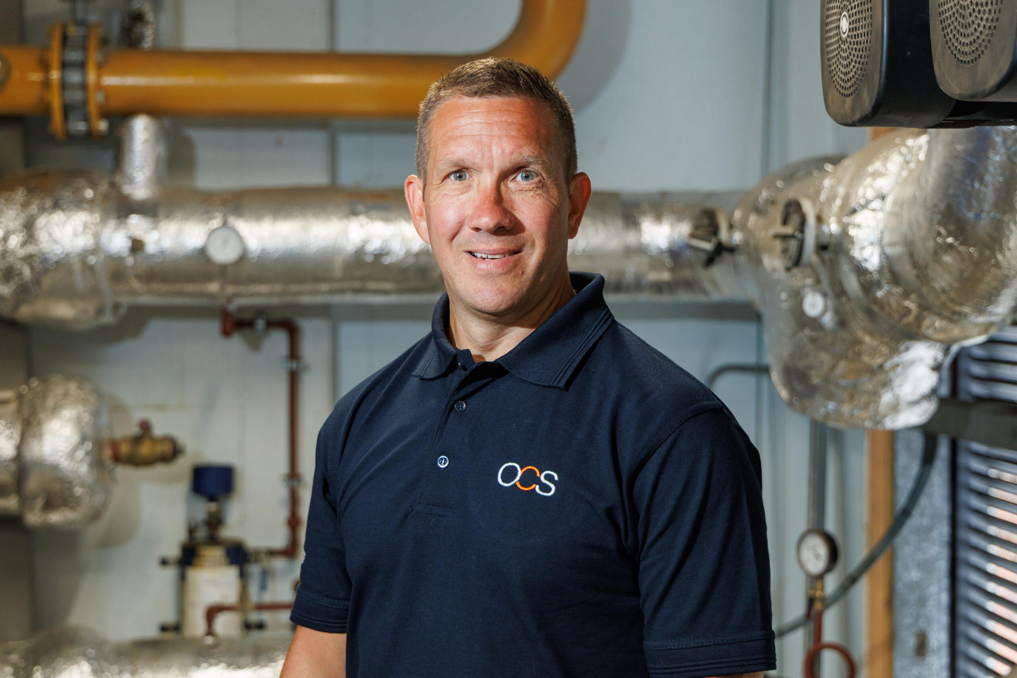 A man in a navy blue OCS-branded polo shirt stands smiling in front of insulated pipes and industrial equipment in a mechanical room.