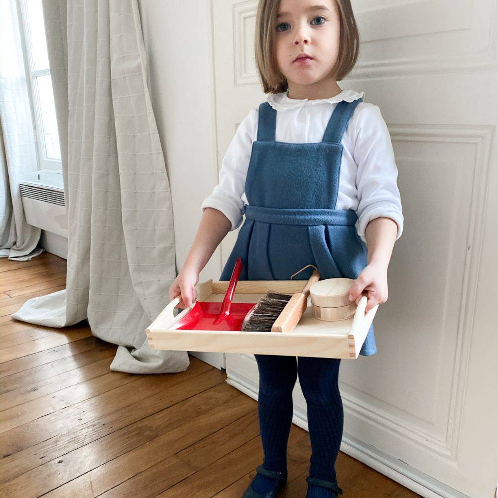 Une petite fille tient un plateau Montessori dans lequelle se trouve une pelle et une balayette
