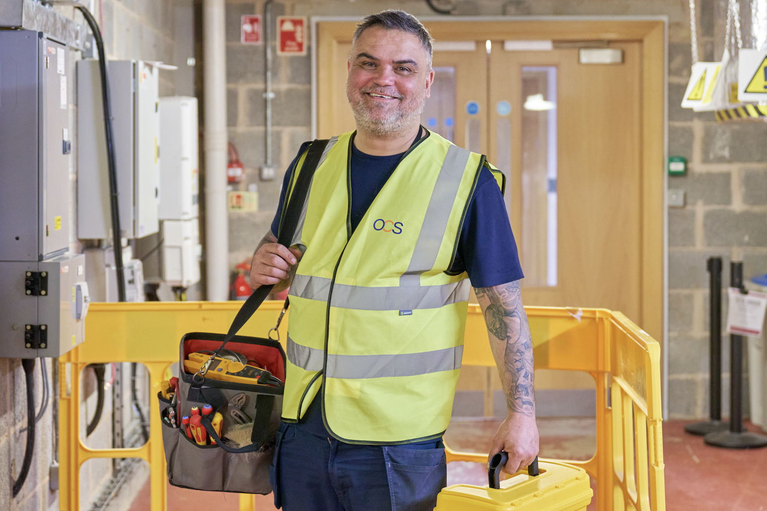 A smiling maintenance worker wearing a high-visibility vest stands indoors, holding a toolbox and a tool bag, surrounded by safety barriers and electrical panels.