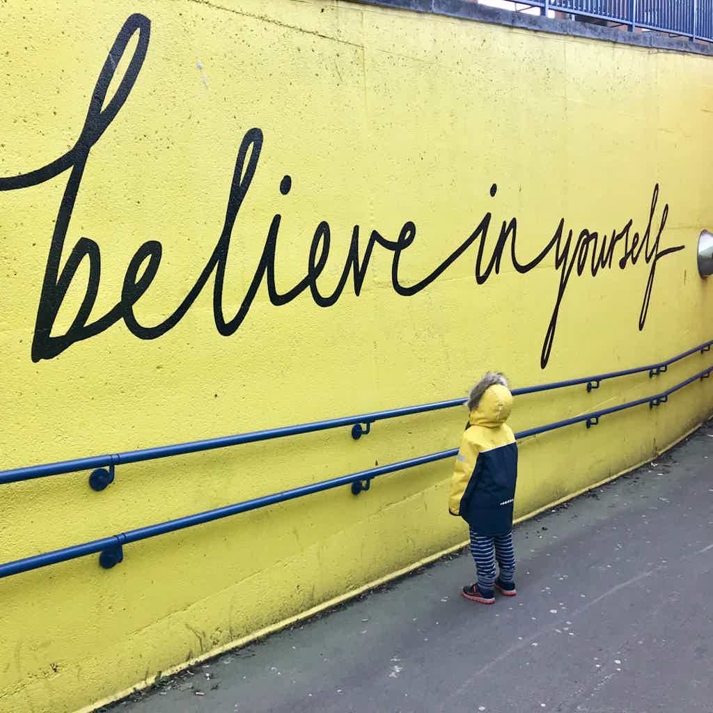 Child in yellow and navy coat stands before a yellow wall with large black cursive text "believe in yourself."
