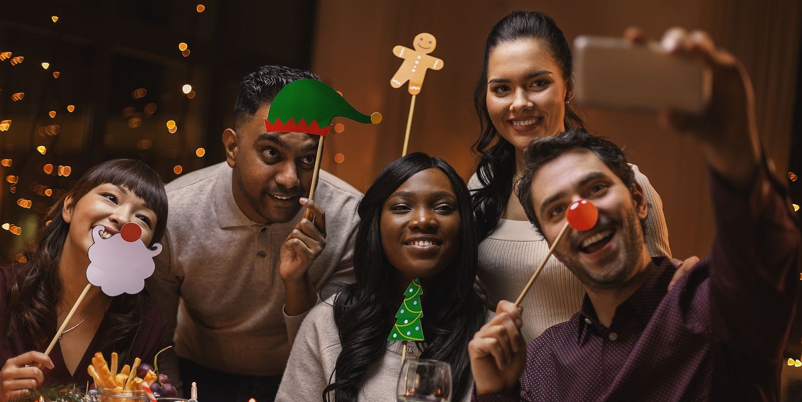 5 friends of different ethnicities sitting at a diner table posing for a selfie with holiday ornaments and smiling.