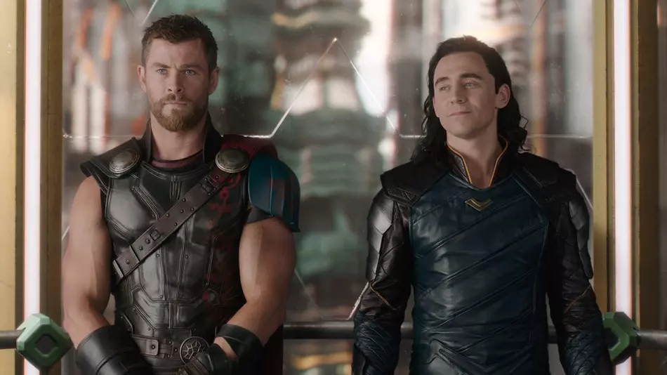 Thor and Loki standing side by side looking intently at someone off camera. Loki smiles as he looks to the side.