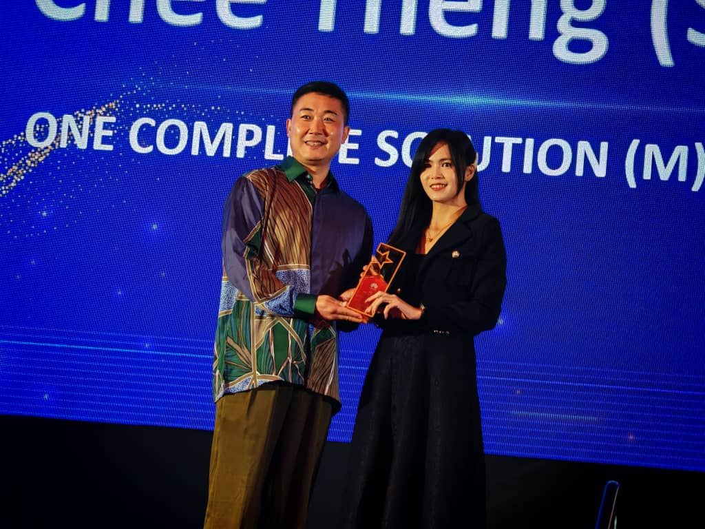 A man and a woman stand on stage smiling, holding an award together. The background displays blue lighting and partially visible text. Both are dressed in formal attire.